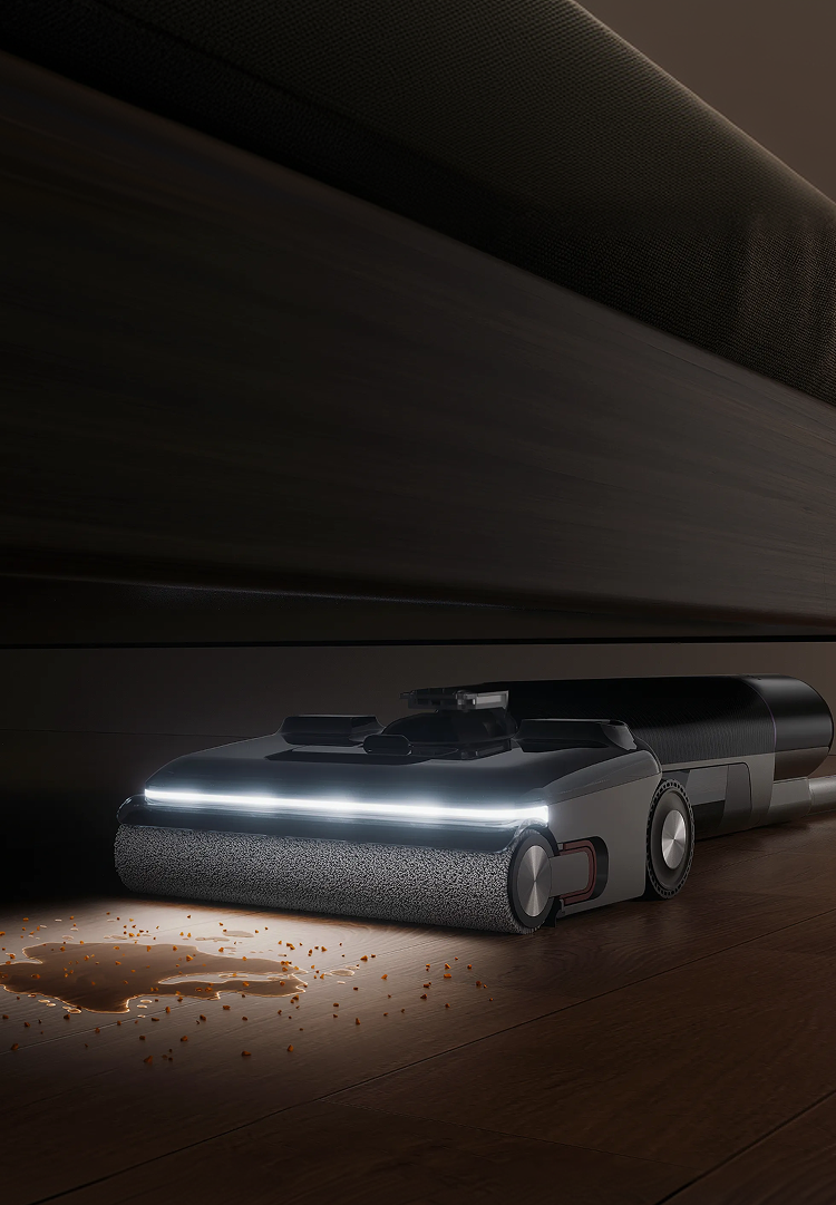A futuristic robotic vacuum cleaner with a textured dark gray body and a bright white front light, shown cleaning under a dark wooden bed frame on a wood floor.